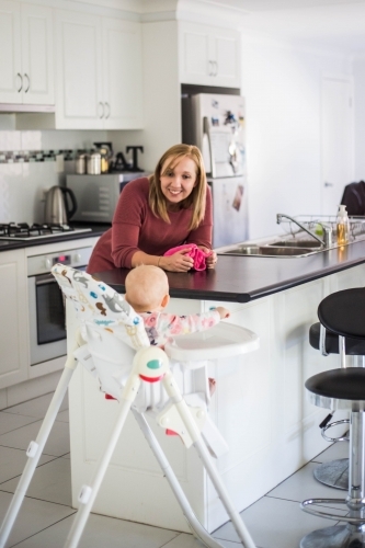 Mother leaning on bench in kitchen smiling at baby in high chair - Australian Stock Image