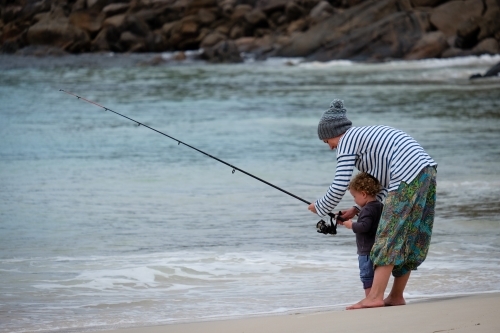 Mother leaning over toddler fishing off beach with rod in cool conditions - Australian Stock Image