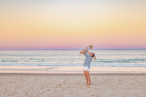Mother lifting toddler son in the air on the beach with pink sunset sky - Australian Stock Image