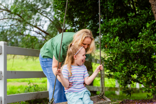 Mother pushing Six year old child on swing in country hanging from jacaranda tree - Australian Stock Image