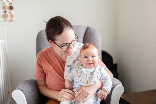 Mother sitting in rocking chair in nursery with delighted baby smiling at the camera - Australian Stock Image