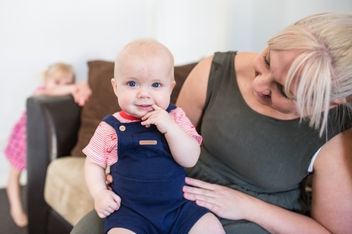 Mother sitting with baby boy on lap with finger in mouth smiling - Australian Stock Image