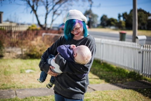 Mother smiling down at child in arms standing in yard with white fence - Australian Stock Image