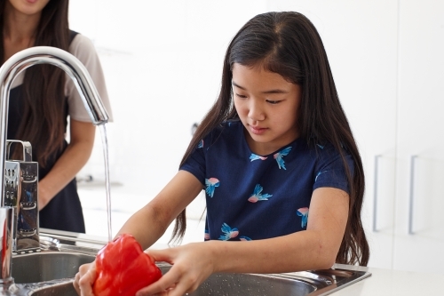 Mother watching over daughter whilst washing vegetable in sink - Australian Stock Image