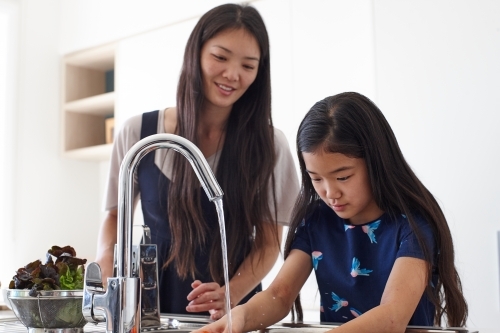 Mother watching over daughter whilst washing vegetable in sink - Australian Stock Image