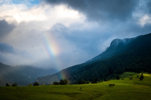 Mountain range covered by clouds with sunlight and rainbow - Australian Stock Image