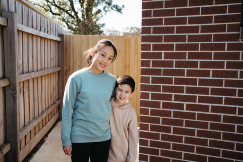 Mum and son standing outside in front of the brick wall of their house. - Australian Stock Image