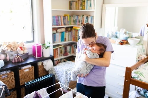 Mum kissing newborn baby standing in messy baby's room - Australian Stock Image