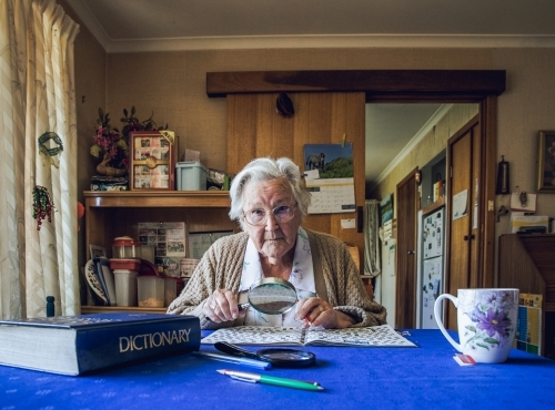 Old lady at dining table with crossword, magnifying glass, dictionary and cup of tea - Australian Stock Image