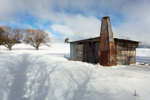 Old timber shack, shed or stable in a rural paddock with full coverage snowfall in winter. Blue sky - Australian Stock Image