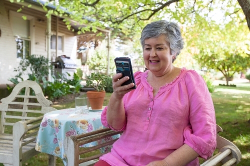 Old woman sitting in backyard looking at her mobile phone - Australian Stock Image