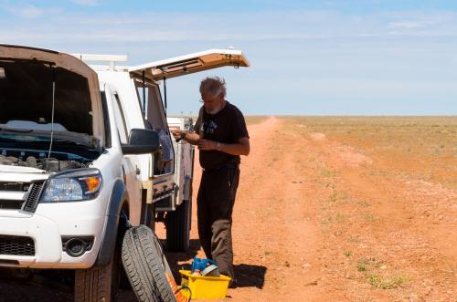 Older grey haired man with beard changing wheel of single cab 4wd with trailer on desert road. - Australian Stock Image