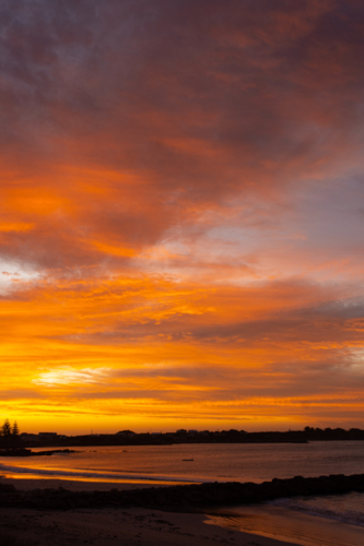 Orange, dramatic sunset over beach and coastal town - Australian Stock Image