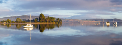 Panorama view of boats reflected on a calm coastal lagoon with lingering fog - Australian Stock Image