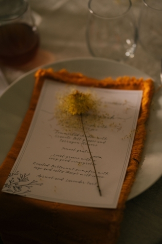 Paper menu on top of the orange napkin with dried yellow flower resting on it
