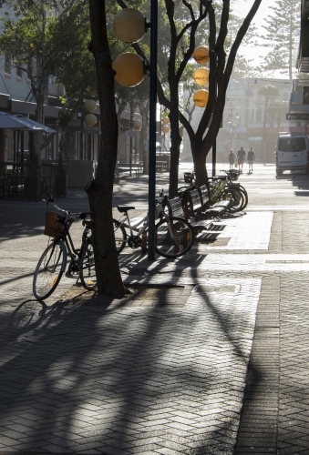 Paved urban mall in early morning light with trees, shadows, parked bicycles and distant pedestrians - Australian Stock Image