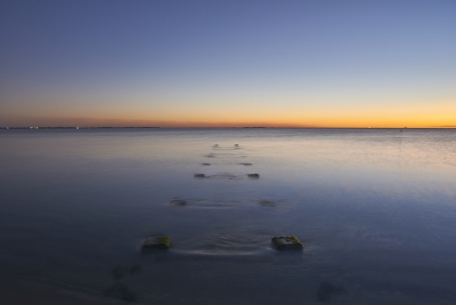 Peaceful ocean at dusk with jetty ruins protruding from the still water. - Australian Stock Image