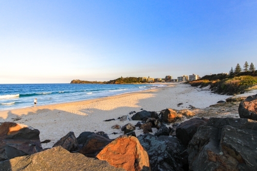 Person walking along sandy beach and rocky coastline beneath blue sky - Australian Stock Image