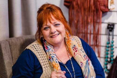 Portrait of a happy middle aged redhead woman sitting in chair at back of clothes shop - Australian Stock Image