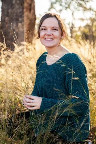 Portrait of a happy young adult in green jumper siting in backlit grass - Australian Stock Image