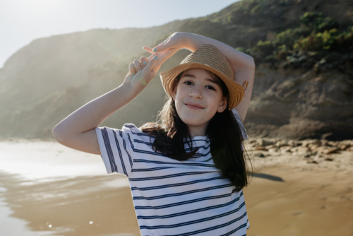 Portrait of a happy young girl wearing a sunhat looking at the camera at the beach in the afternoon - Australian Stock Image