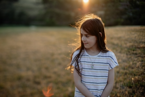 Portrait of a happy young girl with long brown hair standing in a field at sunset - Australian Stock Image