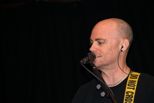 Portrait of a male entertainer singing at microphone with shaved head and black background - Australian Stock Image