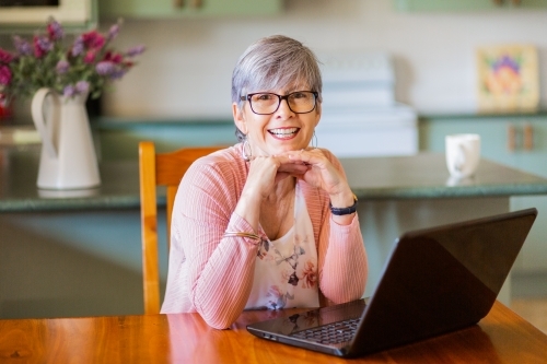 Portrait of a senior woman with glasses sitting in front of laptop inside her home - Australian Stock Image