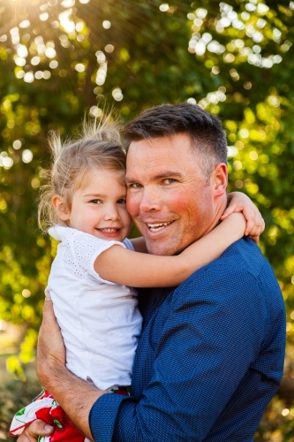 Portrait of a smiling father hugging his happy daughter in golden sunlight - Australian Stock Image