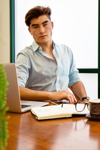 Portrait of a young male office worker sitting with laptop and note book at a meeting table