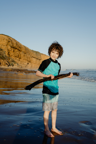 Portrait of happy young boy with curly hair holding a stick on a beach looking at the camera - Australian Stock Image