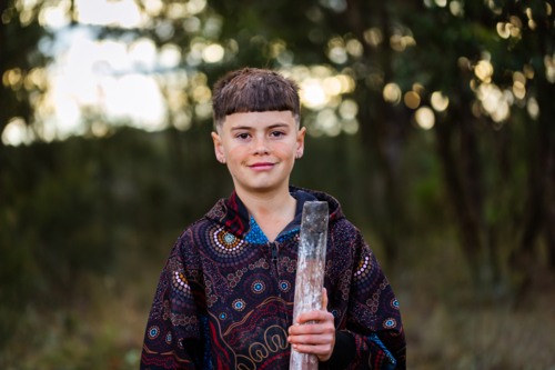 Portrait of young aboriginal boy holding didgeridoo outside with bokeh background - Australian Stock Image