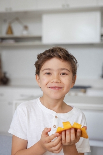 Portrait of young boy eating slice of mango in kitchen - Australian Stock Image