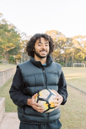 Portrait shot of a young man with curly hair standing on the field holding a soccer ball - Australian Stock Image