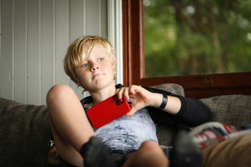 Pre-teen boy sitting on couch holding red mobile phone - Australian Stock Image