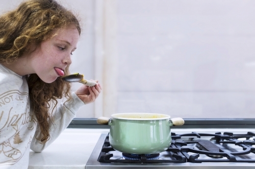 Preteen girl helping to cook over a saucepan on a stove in the kitchen - Australian Stock Image