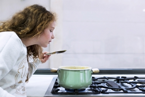 Preteen girl helping to cook over a saucepan on a stove in the kitchen - Australian Stock Image