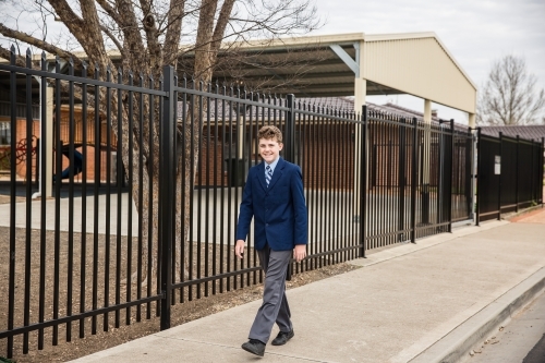 Primary school boy walking on path in front of school near security fence - Australian Stock Image