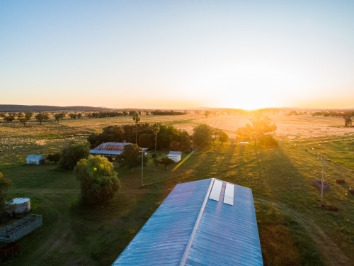Rays of setting sun touching solar panels on farm shed roof, trees casting shadows over homestead - Australian Stock Image