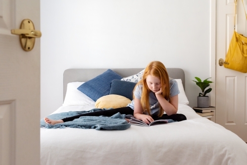 Red haired girl on white bed reading - Australian Stock Image