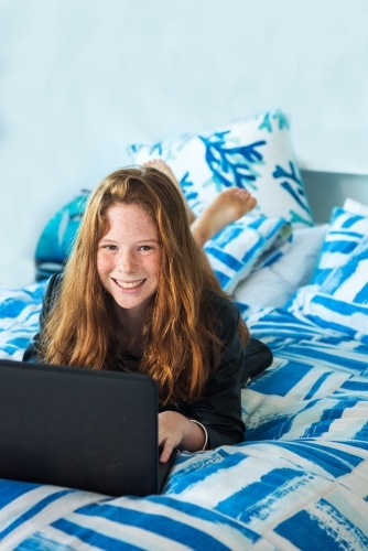 Red haired teenager working on the computer while lying on the bed. - Australian Stock Image