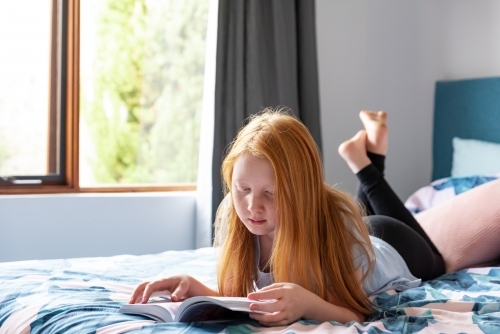 Redhaired girl reading on bed at home - Australian Stock Image