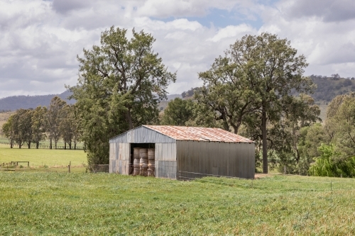 Rural scene with hay bales stacked in an old barn with trees & hills in the background - Australian Stock Image