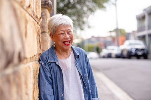 Senior Asian woman laughing while leaning against the wall at the roadside. - Australian Stock Image