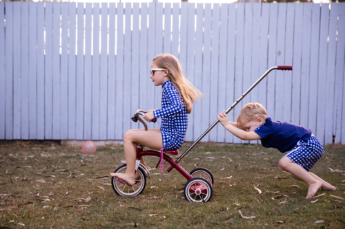 siblings playing on a trike in the back yard with a girl riding and a boy pushing - Australian Stock Image