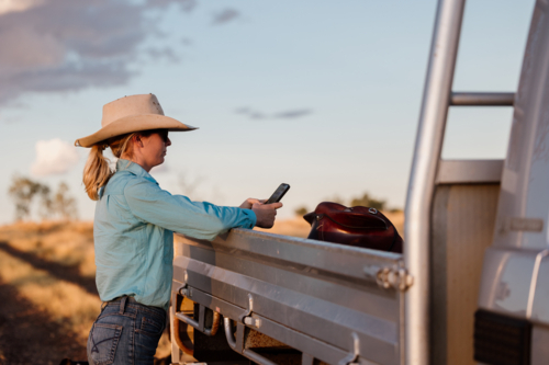 Side profile of female standing looking at a phone leaning on vehicle outdoors in country - Australian Stock Image