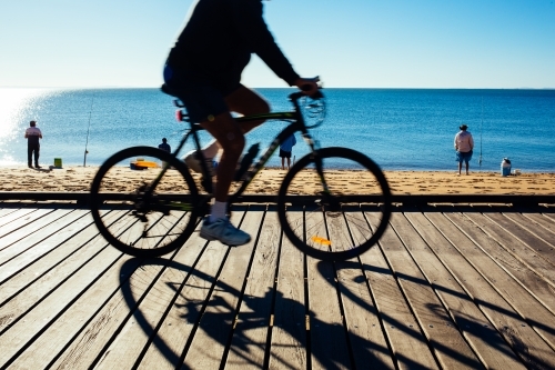 Silhouette of a cyclist riding along a timber boardwalk by the beach at Redcliffe, Queensland. - Australian Stock Image