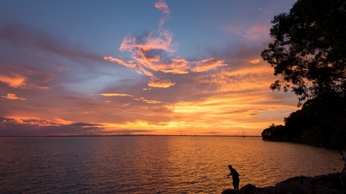 Silhouette of a person standing on a rock at the beach under the sunset sky. - Australian Stock Image