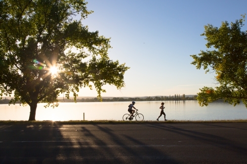 Silhouette of a woman running and a man cycling beside Lake Burley Griffin - Australian Stock Image