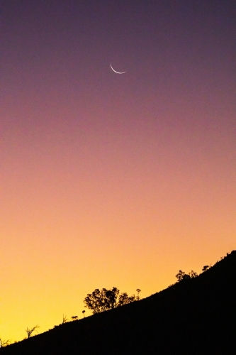 silhouette of hillside against dusk sky with crescent moon - Australian Stock Image